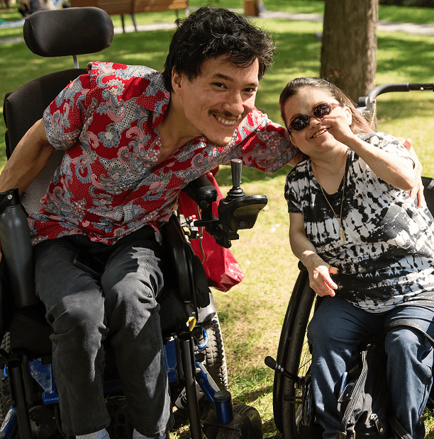 Two people smiling in wheelchairs outdoors.